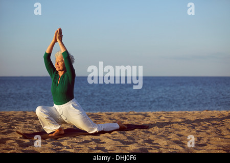 Senior woman practicing yoga on beach. Elderly woman doing yoga exercise on sandy beach. Yoga and relaxation concept. Stock Photo