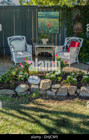Two white garden chairs are placed next to a table and a painting hung on a fence with a cluster of rocks circling a flower bed. Stock Photo
