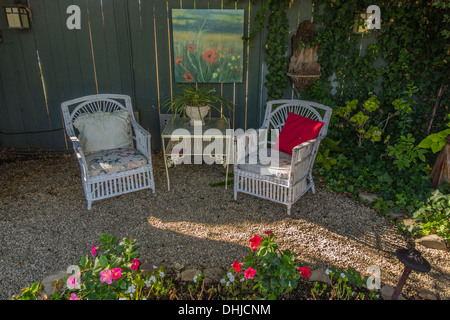 Two white garden chairs are placed next to a table and a painting hung on a fence with a cluster of rocks circling a flower bed. Stock Photo