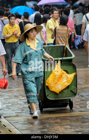 Uniformed young female Asian street cleaner pulling a wheelie bin. Stock Photo