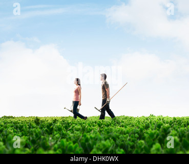 Two young people carrying gardening equipment walking across a green field with plants Stock Photo