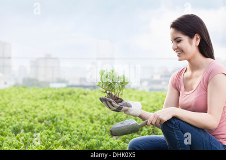 Young smiling woman gardening and holding a plant in a roof top garden in the city Stock Photo