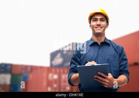 Smiling young engineer in protective work wear in a shipping yard examining cargo Stock Photo