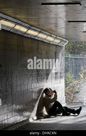 A lone man smoking sits in sunlight at an underpass and leans against a tiled wall to contemplate. Stock Photo