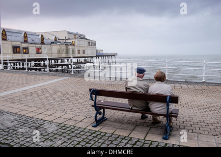 An elderly couple wearing beige grey coats, sitting on a seaside bench on an overcast day Aberystwyth,October 17 2013 Stock Photo