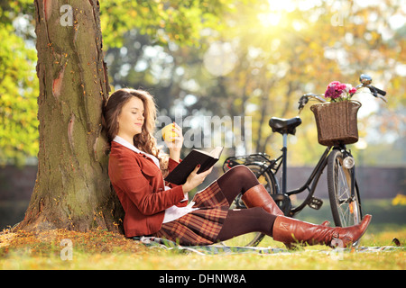 Young female in park reading a book and eating apple, on a sunny day Stock Photo