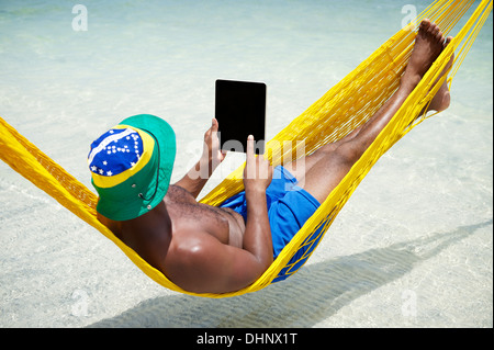 Brazilian man uses tablet computer relaxing in hammock on beach over the sea Stock Photo