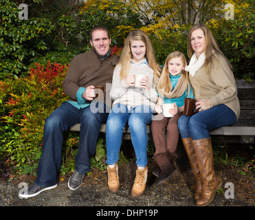 Photo of dad, daughters, and mom all sitting on park bench while taking a break from their walk in the park during a nice autumn Stock Photo
