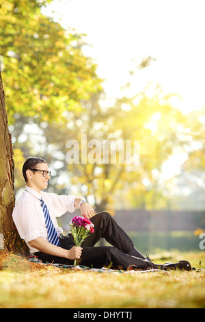 Handsome guy with flowers sitting on a grass and checking the time, in a park Stock Photo