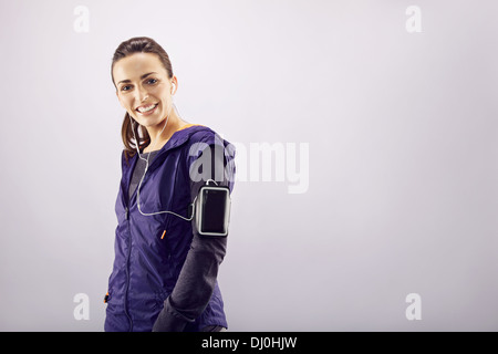 Portrait of cheerful young female runner listening to music while standing against grey background. Caucasian fitness woman Stock Photo