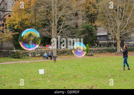 A woman makes two large soap bubbles showing rainbow colours whilst children watch in awe. Betws-y-Coed, Conwy, North Wales, UK Stock Photo