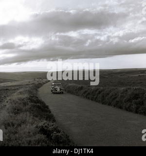 Historical picture from1950s England, showing a car travelling along an empty country lane with hedgerows. Stock Photo