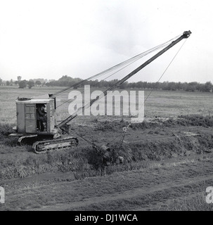 1950s, historical picture of a mechanical dragline bucket on crane with tracks positioned in a country field, excavating earth from a small river or brook that runs beside it, England, UK. Stock Photo