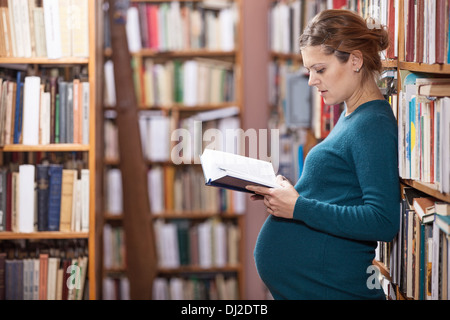 Young pregnant woman reading book in library Stock Photo