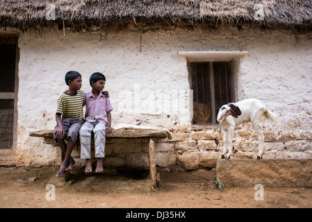 Two indian boys sat outside a thatched rural indian house looking at a goat. Andhra Pradesh, India Stock Photo