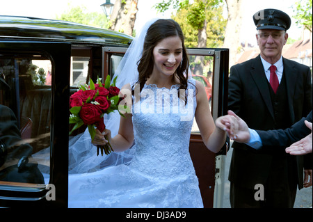 Bride arriving in traditional white wedding dress carrying bouquet of red roses arriving at church with her chauffeur vintage car and a helping hand Stock Photo
