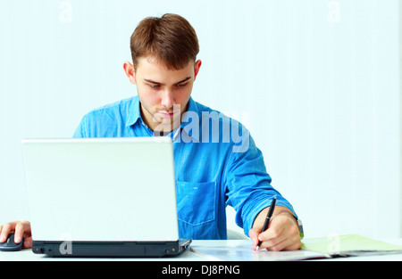 Young concentrated businessman writing notes down with laptop at office Stock Photo