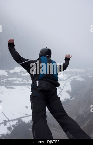 BASE jumper is exiting from a cliff down into the deep valley. Thereby he's keeping his arms up to be stable and save. Stock Photo