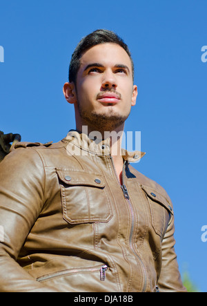 Handsome young man in leather jacket against blue sky looking far, shot from below. Stock Photo