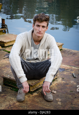 Attractive young man sitting on rusty metal next to river waters, looking at camera. Stock Photo