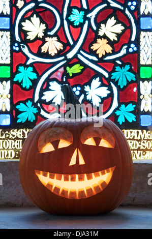 All Hallows Eve. A Halloween Jack o’ Lantern carved from a pumpkin, glowing by candlelight, in front of a stained glass window. England, United Kingdom. Stock Photo