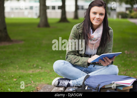 Happy brunette student using tablet sitting on bench Stock Photo