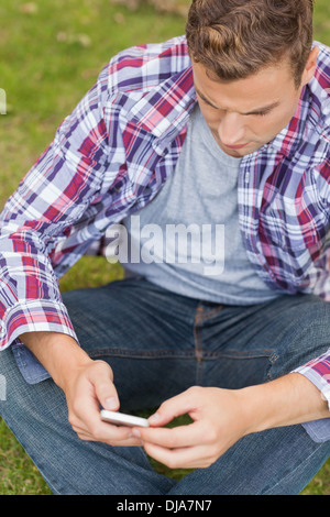 Handsome unsmiling student sitting on grass texting Stock Photo
