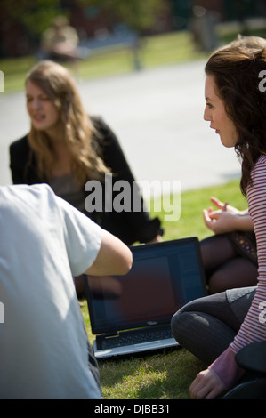 female university students sat on grass with laptop computer talking in sunshine Stock Photo