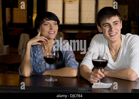 Happy young couple drinking red wine at the bar smiling in amusement as they watch something off screen to the right Stock Photo