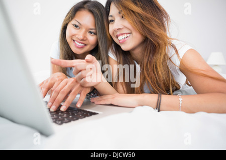 Two smiling brunette sisters lying on a bed using a laptop Stock Photo