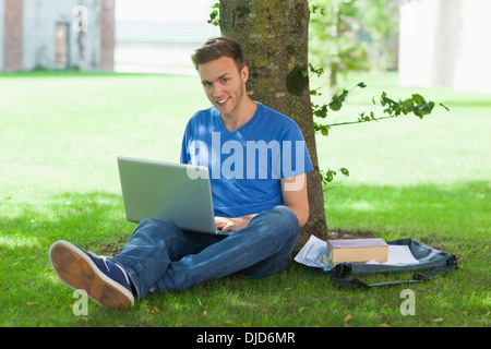 Smiling handsome student sitting under tree using laptop Stock Photo