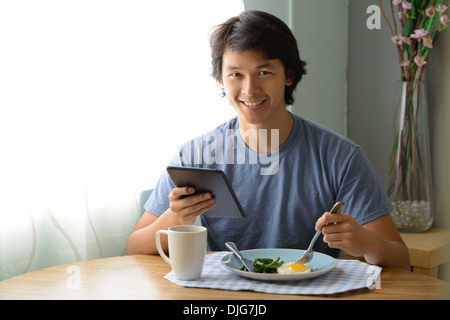 A young Asian man holding an ipad smiling and eating breakfast. Stock Photo