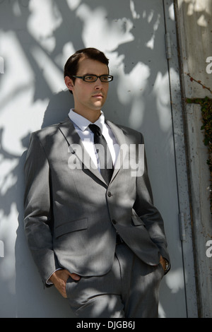 A businessman in gray business suit, wears eye glasses leaning against a gray door, resting in the shadow with hands in pocket. Stock Photo