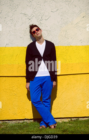 A young man wearing a black sweater, white shirt, and blue pants, standing against a bright neon yellow wall, a fashion concept