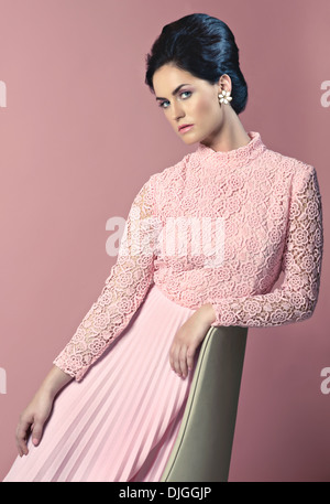 A beautiful woman dressed in a 1960s dress posing on a beige vintage mid century chair, a retro style fashion photograph. Stock Photo