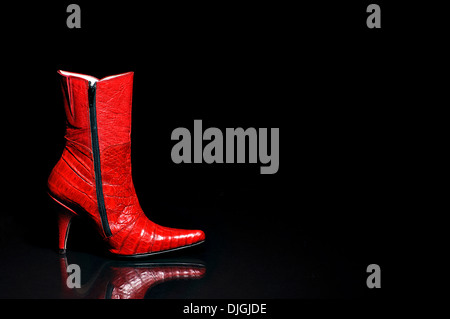 A red crocodile leather boot taken sideways resting on a reflective black surface, with black background. Women's shoes fashion. Stock Photo