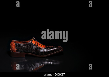 A brown crocodile leather shoe taken sideways resting on a reflective black surface, with black background. Men's shoes fashion. Stock Photo