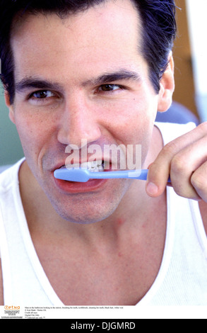 Man in the bathroom looking in the mirror, brushing his teeth, toothbrush, cleaning his teeth, black hair, white vest Stock Photo