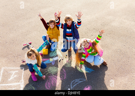 Group of four boys and girls, friends in autumn clothes painting with chalk on the asphalt lifting hands with smile on theirs faces Stock Photo