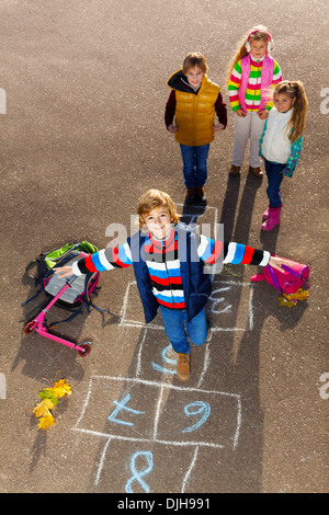 Boy jumping on hopscotch game with friends boys an girls standing by with school bags laying near Stock Photo