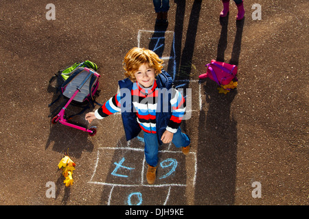 Nice blond boy jumping over hopscotch game after school with bags and scooter laying near Stock Photo