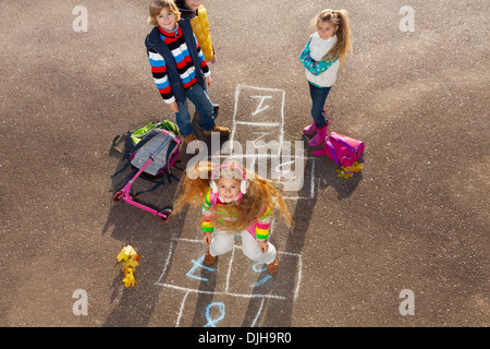Happy girl jumping on hopscotch game with friends boys an girls standing by with school bags laying near Stock Photo
