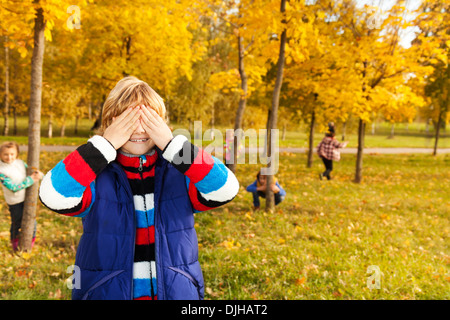 Kids play hide and seek with boy counting covering face with palms while others hiding behind trees Stock Photo