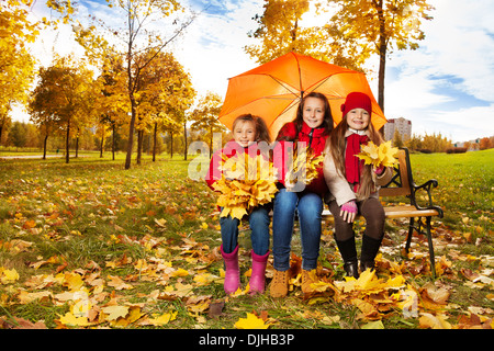 Three happy girls sitting on the bench under the umbrella and with maple bouquet in the autumn park Stock Photo