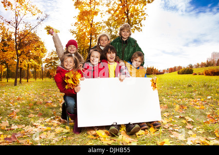 Happy smiling group of kids, friends, boys and girls, showing blank placard board to write it on your own text in autumn park with maple trees Stock Photo