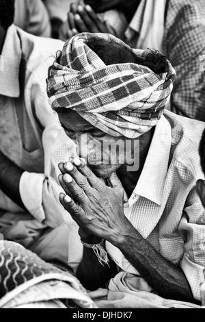 Old Indian man in prayer whilst waiting at Sri Sathya Sai Baba mobile outreach hospital. Andhra Pradesh, India. Monochrome Stock Photo