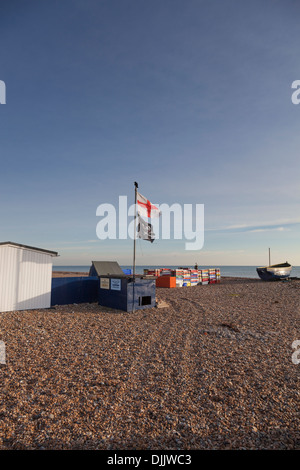 Flag of St George and the 'Jolly Roger' pirate flag fly on a shingle beach amidst fish containers and a fishing boat. Stock Photo