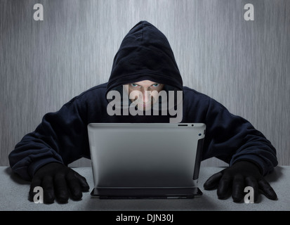 A hooded male representing a cyber criminal, looking at a tablet computer. Stock Photo