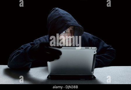 A hooded male representing a cyber criminal, using a tablet computer. Stock Photo