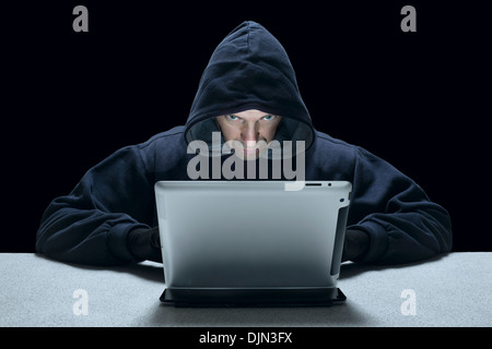 A hooded man representing a cyber criminal, using a tablet computer. Stock Photo